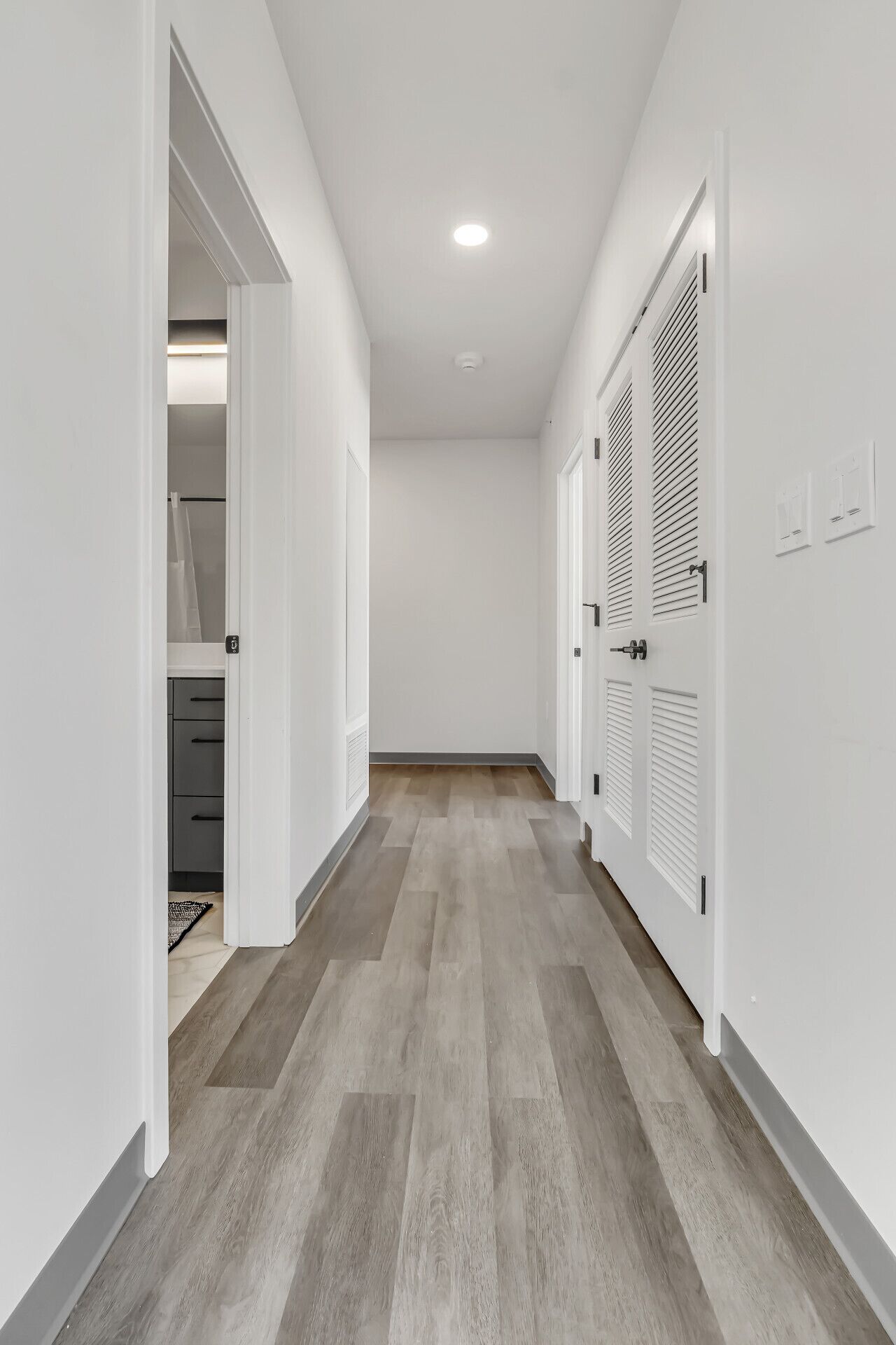 A narrow hallway with wooden flooring, white walls, and doors with louvered shutters.