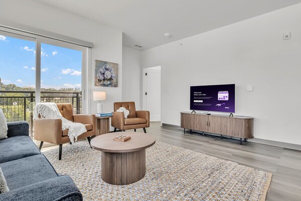 A modern living room with a large TV, comfortable seating, and a view through large windows.