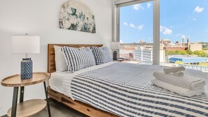 Een slaapkamer met een groot raam, een bed met gestreept beddengoed, een nachtkastje met een lamp, en uitzicht op gebouwen.