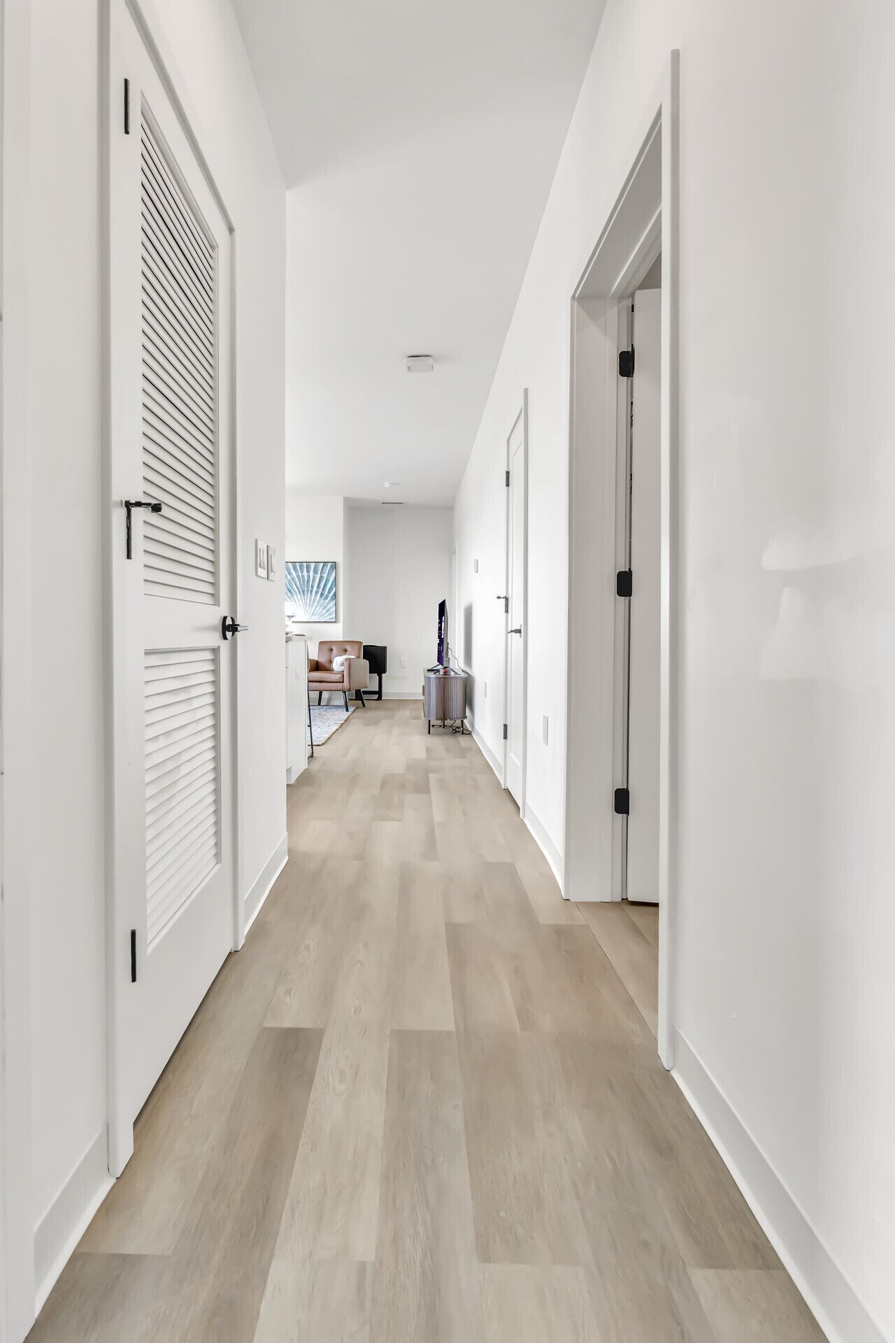 A modern, well-lit hallway with white walls and wooden flooring, leading to a kitchen area with a dining table and chairs.