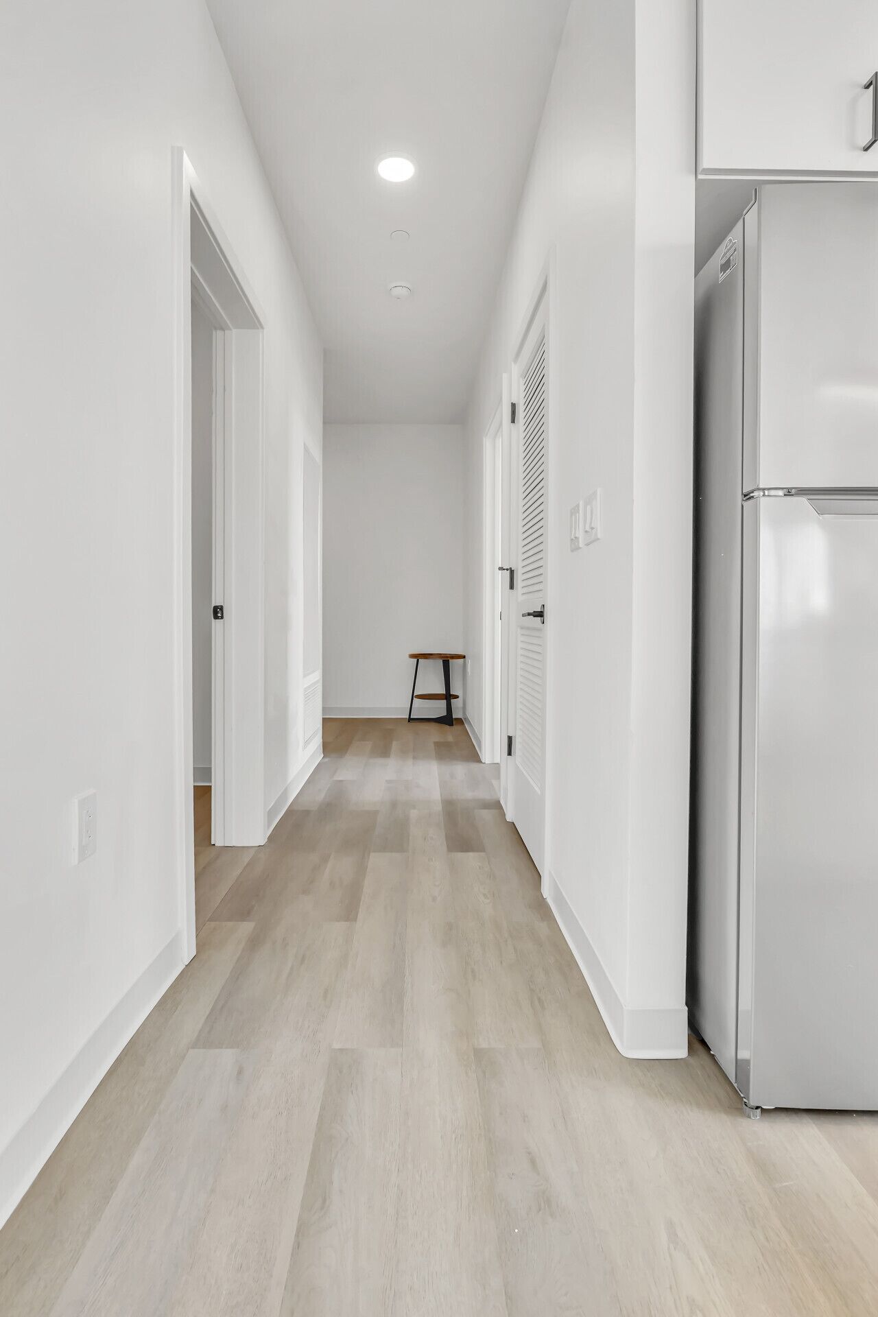 Un pasillo estrecho con suelo de madera, paredes blancas y un refrigerador a la derecha.