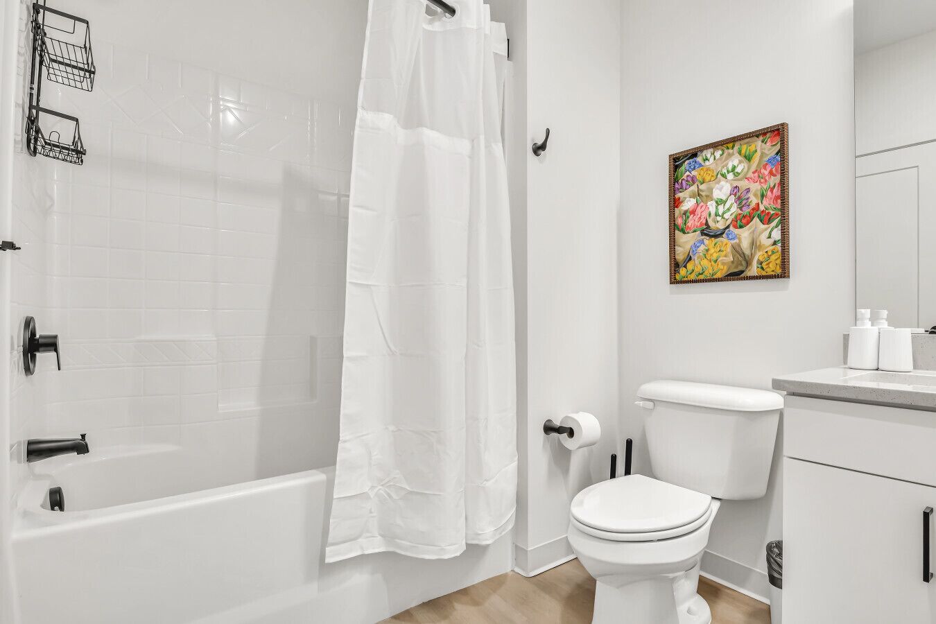 Une salle de bain avec une baignoire blanche, des toilettes et un rideau de douche. Il y a un tableau au mur et un lavabo avec des articles de toilette.