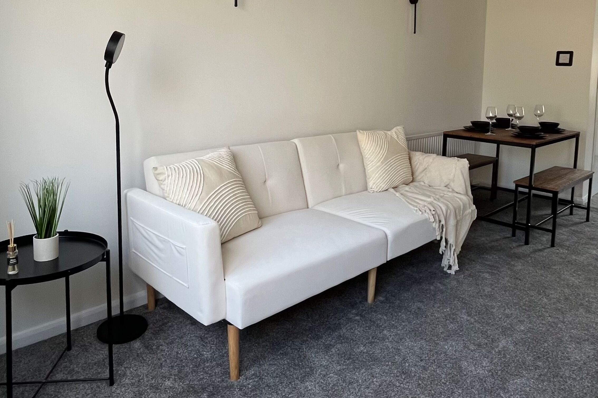 Un canapé blanc avec un plaid, une petite table ronde ornée d’une plante et un lampadaire.
