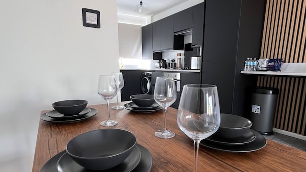 A modern kitchen with dark cabinetry, a wooden dining table set with black plates and wine glasses, and a coffee machine. - 2 bed House in City Centre - Offers for Long Stays (Nottingham)