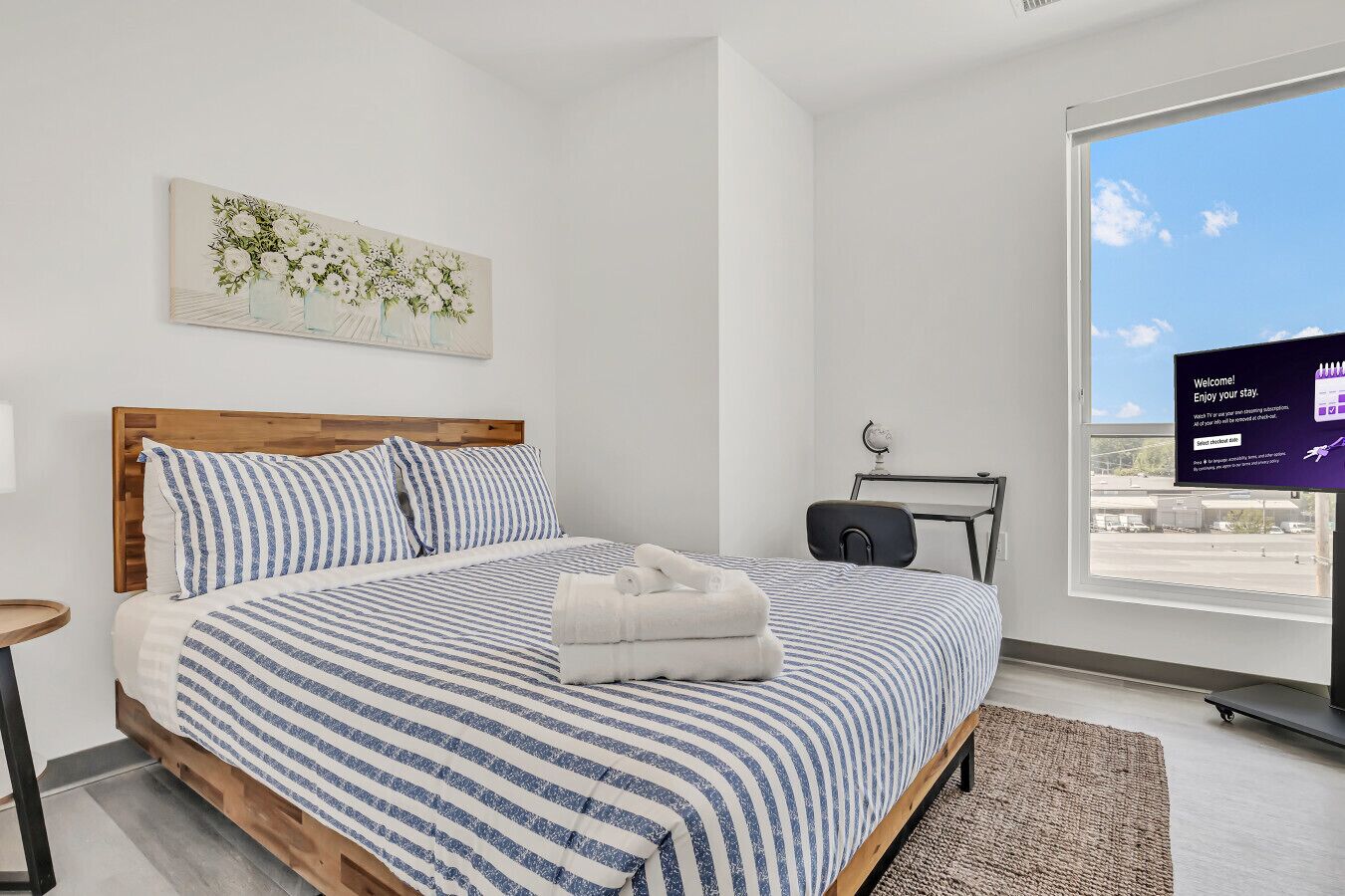 A modern bedroom with a wooden bed, striped bedding, a small bedside table, a globe, and a window with a view of the sky.