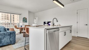 Une cuisine moderne équipée d’un lave-vaisselle, un coin repas avec une table et des chaises, et une vue sur la ville grâce à de grandes fenêtres.