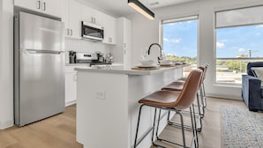 Une cuisine moderne dotée d’appareils électroménagers en acier inoxydable, d’un îlot central avec des tabourets de bar et de grandes fenêtres offrant une vue sur la ville.