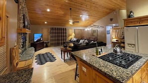 A spacious living area with a fireplace, a stone wall, a large window with curtains, a sofa, a dining table, and a kitchen with granite countertops and a stove.