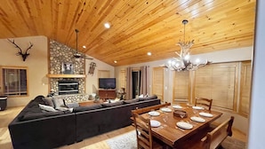 A cozy living room with a wooden ceiling, a stone fireplace, a large sectional sofa, and a dining area with a wooden table set for four.