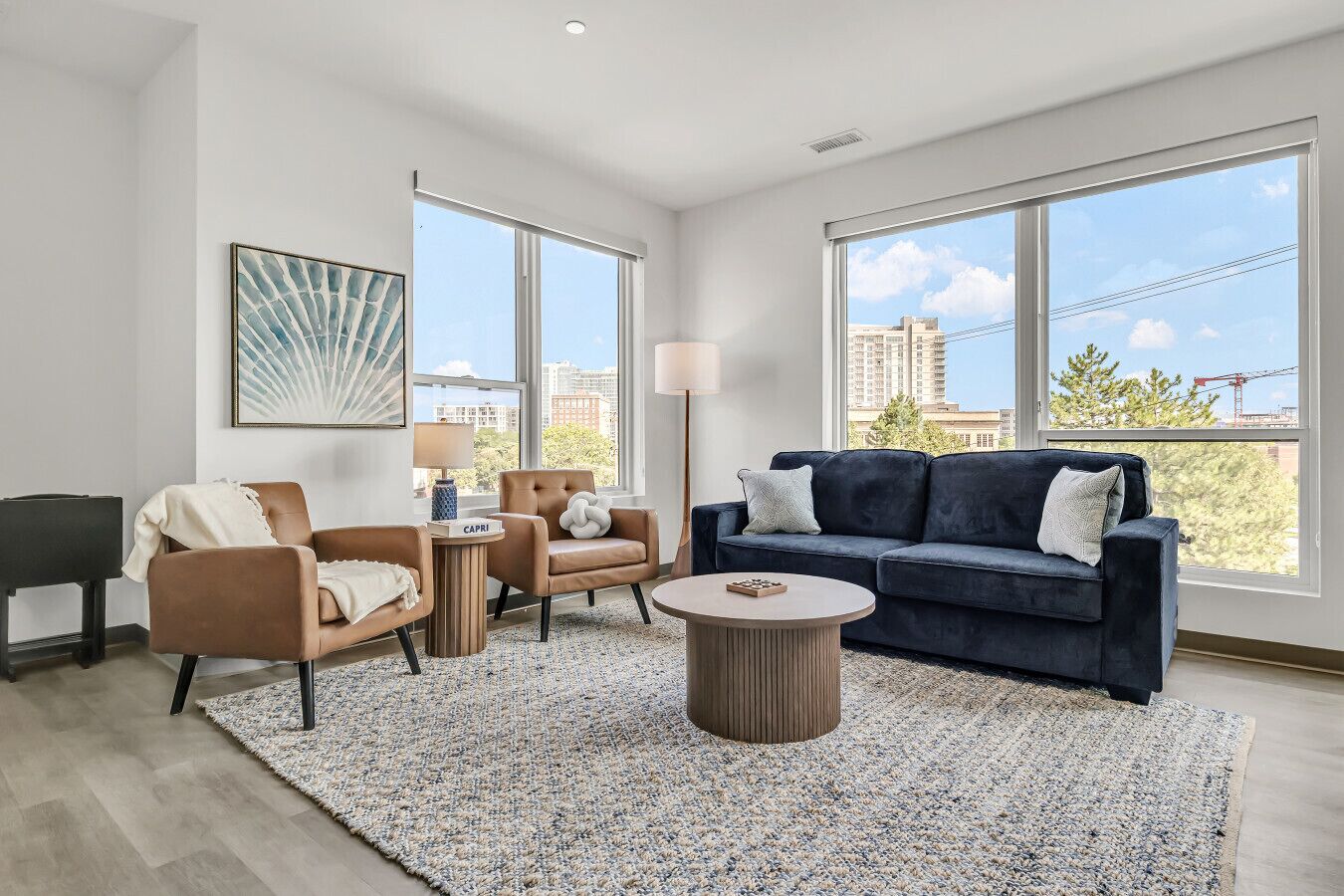 A modern living room with a dark blue sofa, a brown leather armchair, a round coffee table, and a piano. Large windows offer a view of the cityscape.