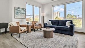 Uma sala de estar moderna com um sofá azul escuro, uma poltrona de couro marrom, uma mesa de centro redonda e um piano. Grandes janelas oferecem vista para a paisagem urbana.