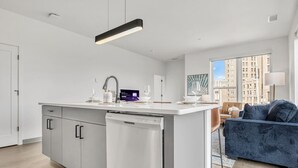 Cozinha moderna com máquina de lavar louça, área de refeições com sofá e vista para edifícios através de grandes janelas.