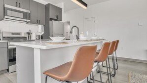 Uma cozinha moderna com armários brancos, uma ilha central com banquetas, eletrodomésticos de aço inoxidável e uma luminária pendente.