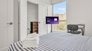 Quarto de hotel com cama, escrivaninha, cadeira, TV e vista para edifícios pela janela.