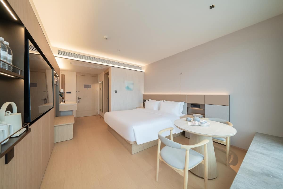 Quarto de hotel moderno com uma cama grande, uma mesa de cabeceira, uma mesa de jantar redonda e uma pequena cozinha equipada.