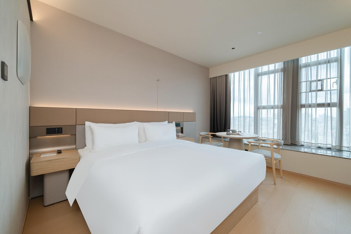 A modern hotel room with a large bed, bedside table, and a small desk with a chair.