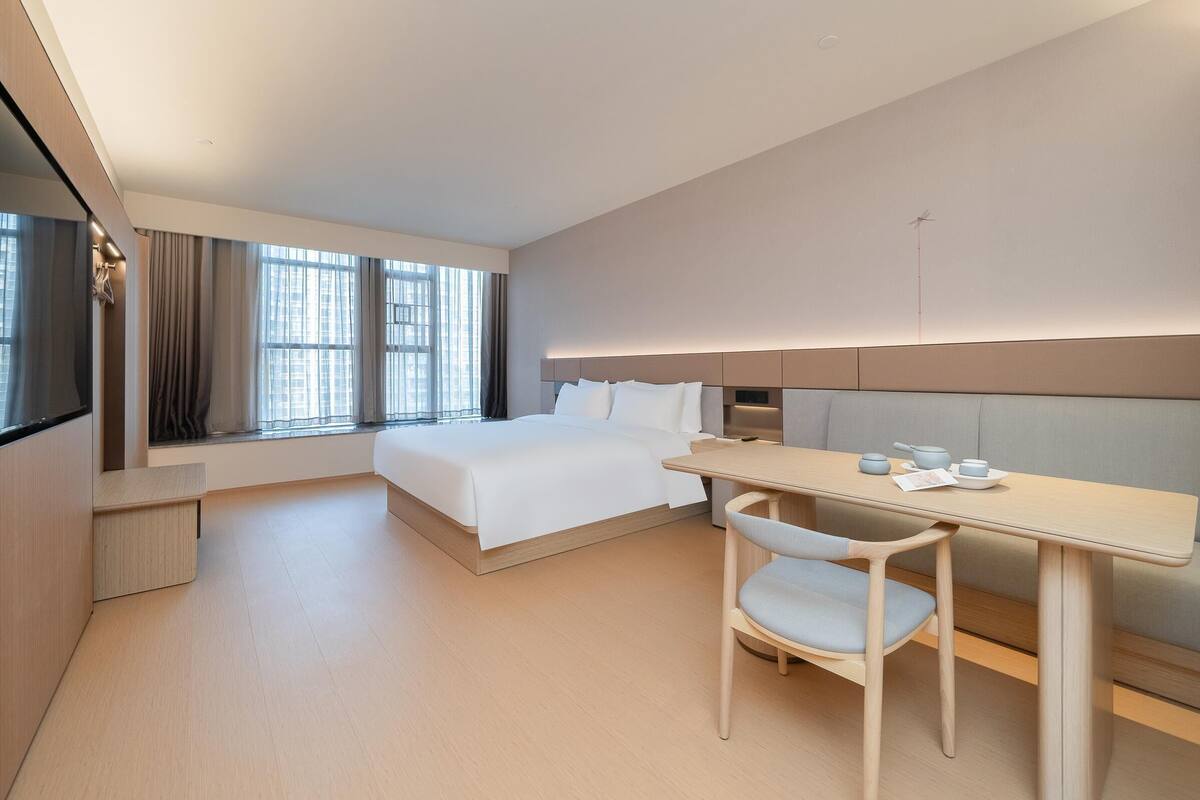 Quarto de hotel moderno com uma cama grande, uma escrivaninha com cadeira e uma janela com cortinas.