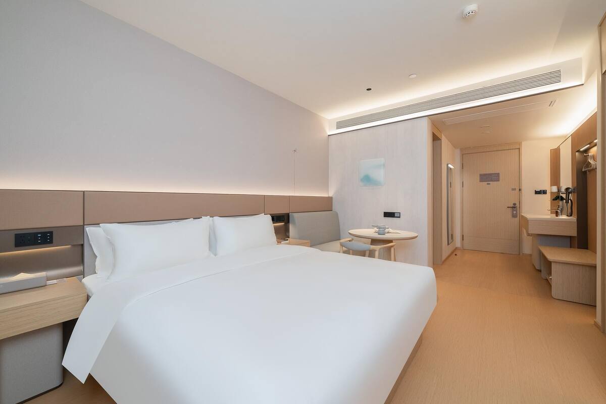 Quarto de hotel moderno com uma cama grande, mesas de cabeceira, uma pequena mesa redonda, um espelho e um banheiro separado.
