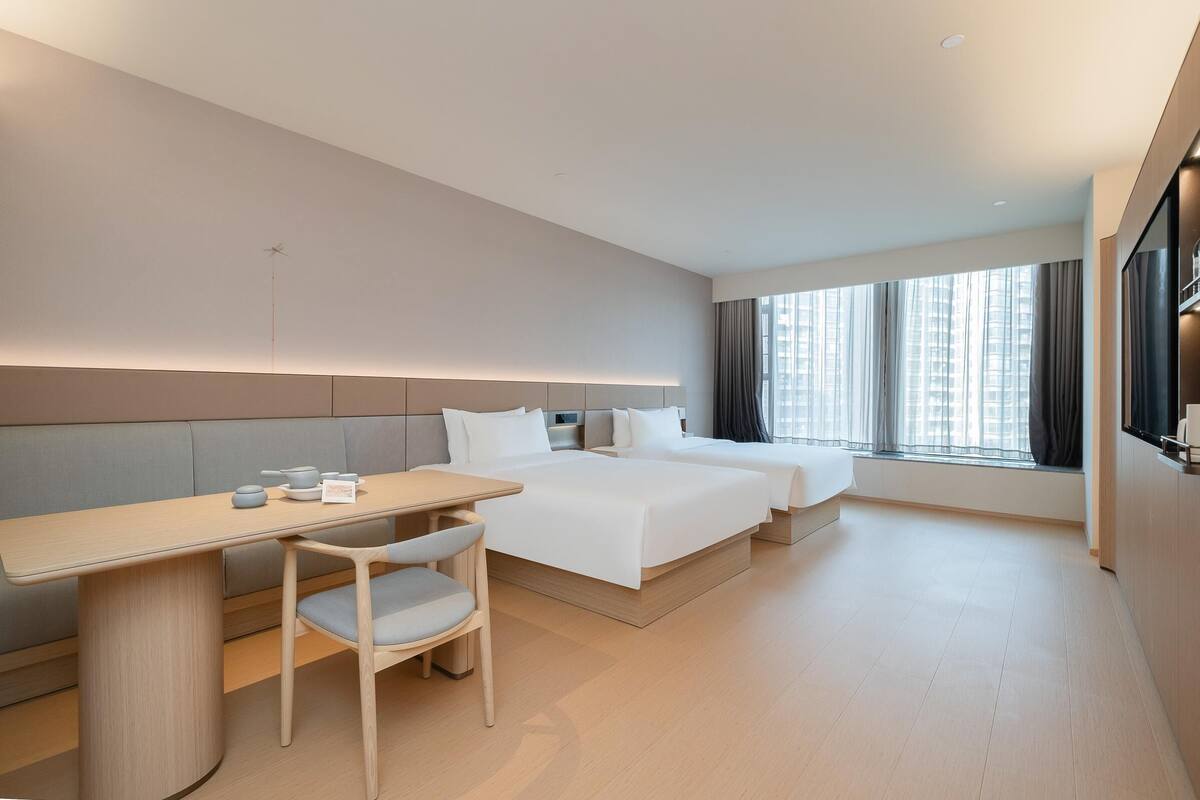Quarto de hotel moderno com uma cama, uma escrivaninha, uma cadeira e uma janela grande.