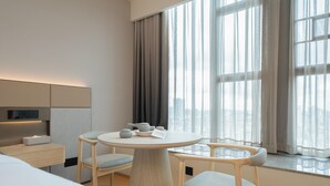 Ein modernes Hotelzimmer mit einem runden Esstisch, zwei Stühlen, einem Bett, einem Nachttisch und einem großen Fenster mit Vorhängen.