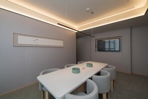Un salón de conferencias con una larga mesa, sillas tapizadas en gris, una obra de arte enmarcada en la pared y una ventana con persianas.