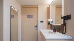 Ein modernes Badezimmer mit Holztür, großem Spiegel, weißem Waschbecken und einem an der Wand befestigten Föhn.