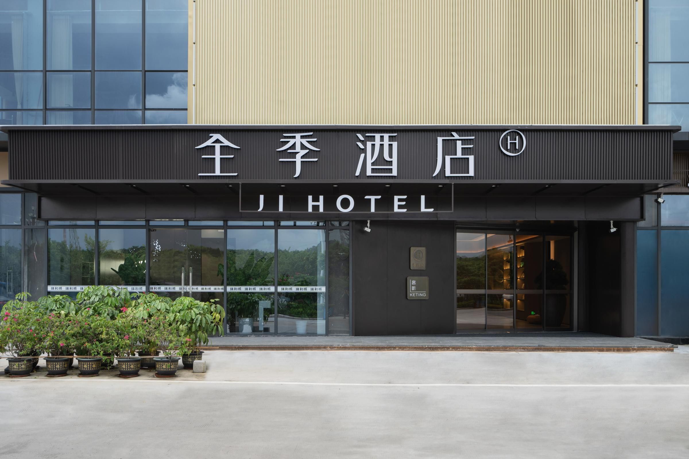 Entrada de hotel com fachada moderna, placa em chinês e inglês e um pequeno jardim com vasos de plantas.