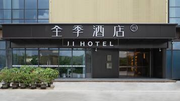 Entrada de hotel com fachada moderna, placa em chinês e inglês e um pequeno jardim com vasos de plantas.