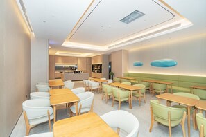 A modern dining area with wooden tables and chairs, a kitchen in the background, and a green cushioned bench along the wall. - Ji Hotel Guangzhou Huangpu Wenchong (Guangzhou)