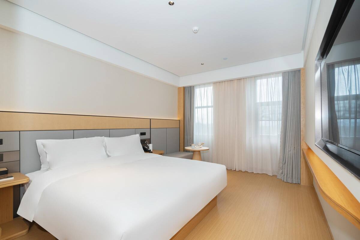 A hotel room with a large bed, a bedside table, a small table with a cup, and a window with curtains.