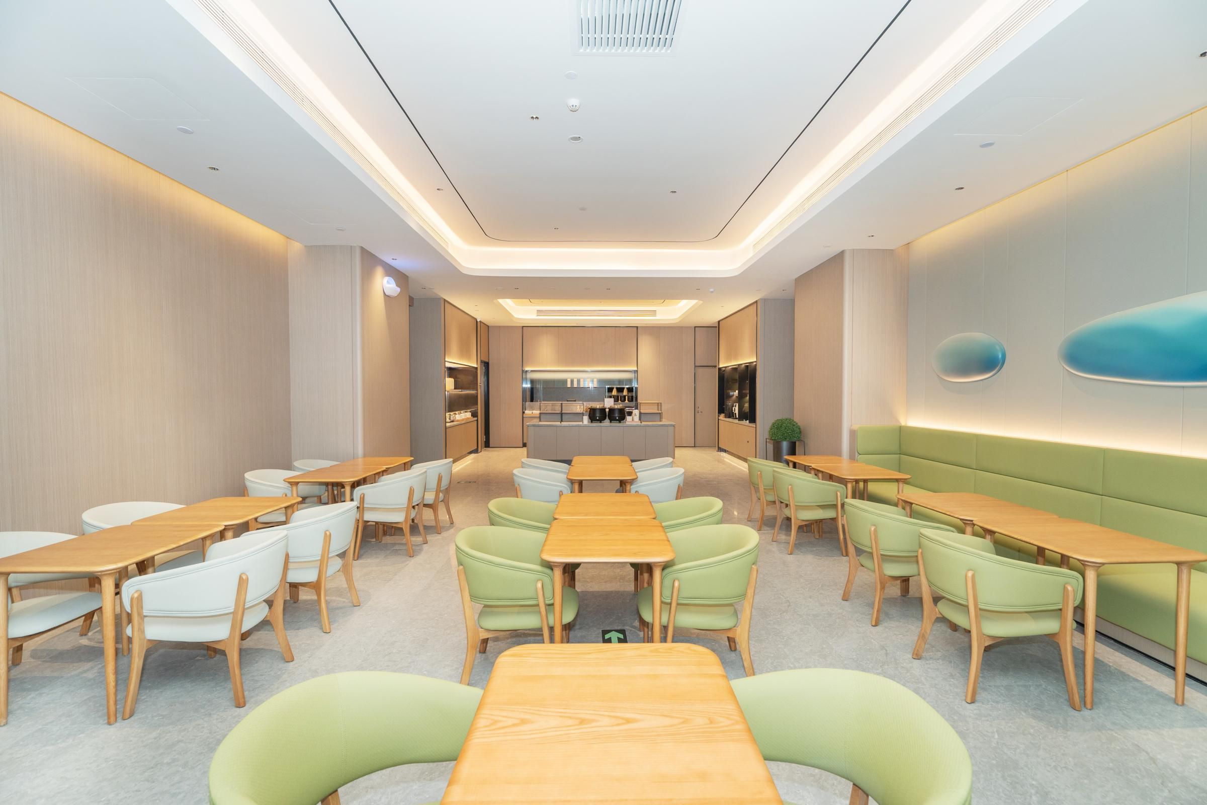 Un comedor moderno con mesas de madera y sillas tapizadas de verde, una zona de cocina central y un área de estar con un sofá verde.