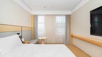 A hotel room with a large bed, a bedside table with a lamp, a small round table with a cup and a book, and a TV mounted on the wall.