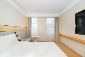 Una camera d'albergo con un letto grande, un comodino con una lampada, un tavolino rotondo con una tazza e un libro, e un televisore montato a parete.