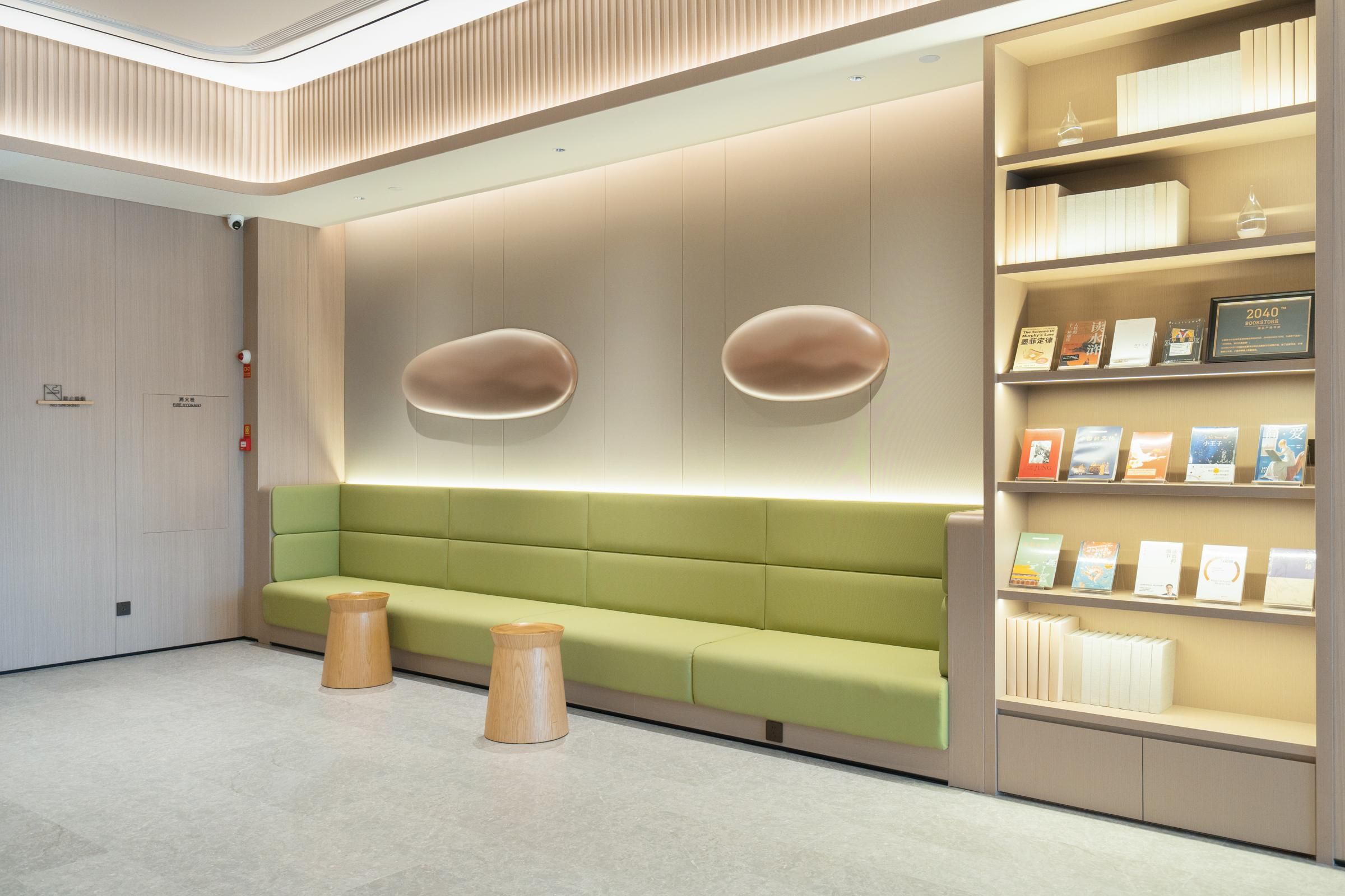 Una zona de espera moderna con un sofá verde, taburetes de madera y una estantería con libros y una placa.