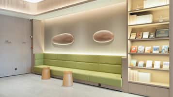 Una zona de espera moderna con un sofá verde, taburetes de madera y una estantería con libros y una placa.