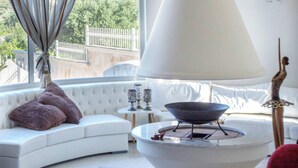 Un salon moderne avec un canapé blanc, un grand vase blanc, une petite table agrémentée d’objets décoratifs et une vue sur un jardin par la fenêtre.