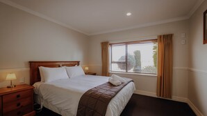 A hotel room with a large bed, a nightstand with a lamp, a window with curtains, and a view of the outside. - Alpine Springs Motel | Unit 8 - HS61218 (Hanmer Springs)