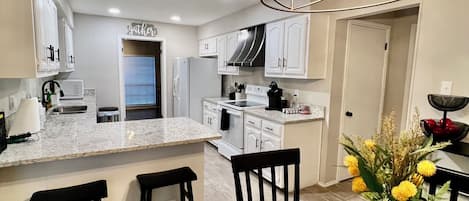 Una cocina moderna con armarios blancos, una isla central y un comedor con mantel a cuadros y un jarrón de flores.