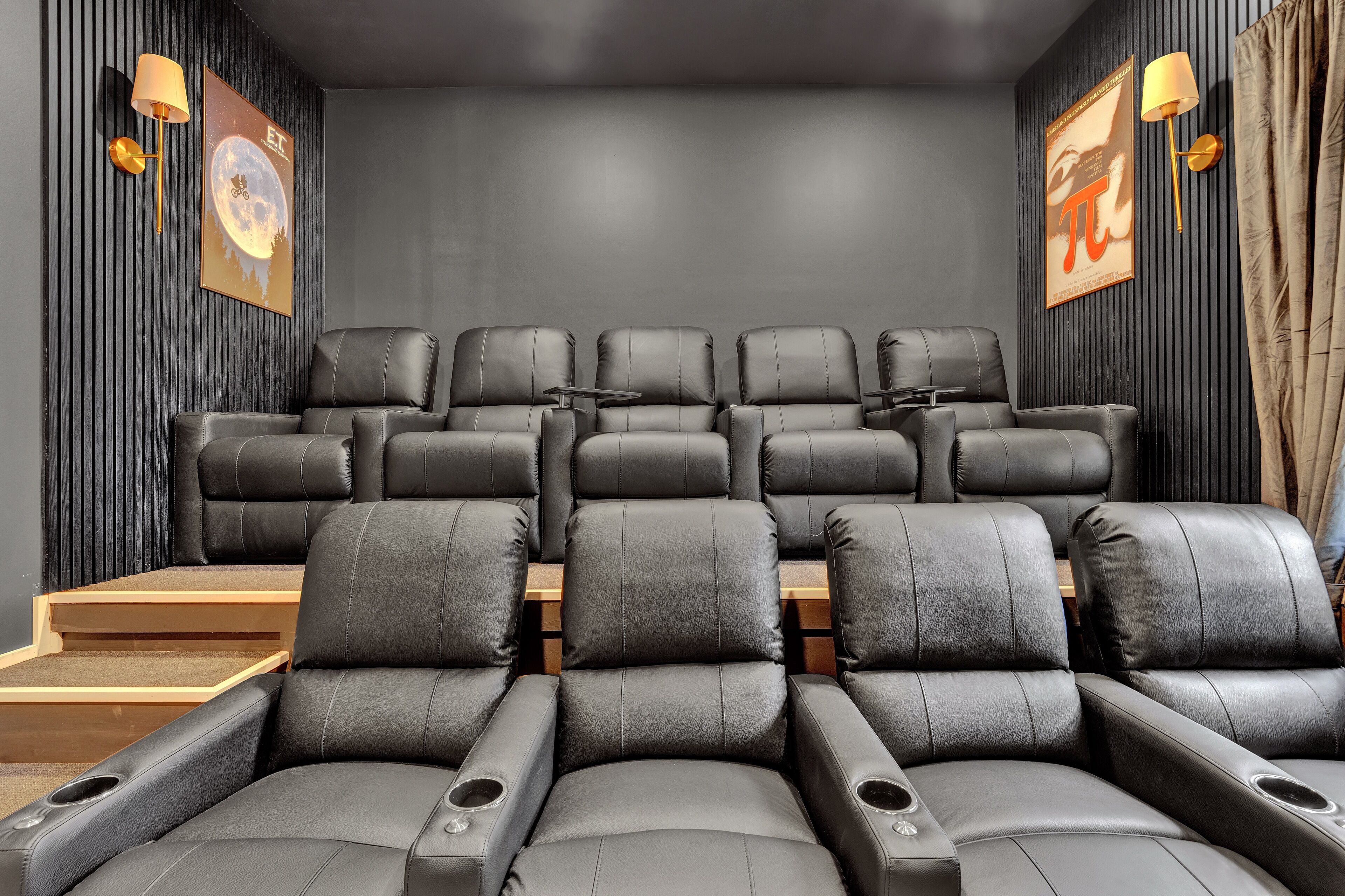 Un cine en casa con varios sillones de cuero negro reclinables, escalones de madera y dos lámparas fijadas a la pared.