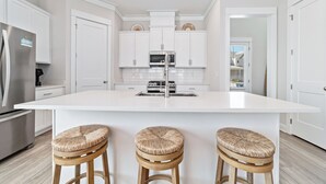 A modern kitchen with white cabinetry, a central island with stools, stainless steel appliances, and a view to the outside. - Salt and Sand at Summer Salt | Zero Entry Pool | Beach Access | Lake Fishing | My Beach Getaways (Orange Beach)