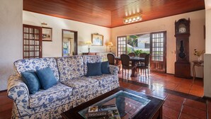 A living room with a floral sofa, a glass coffee table, a dining table with chairs, a wooden ceiling, and a clock.