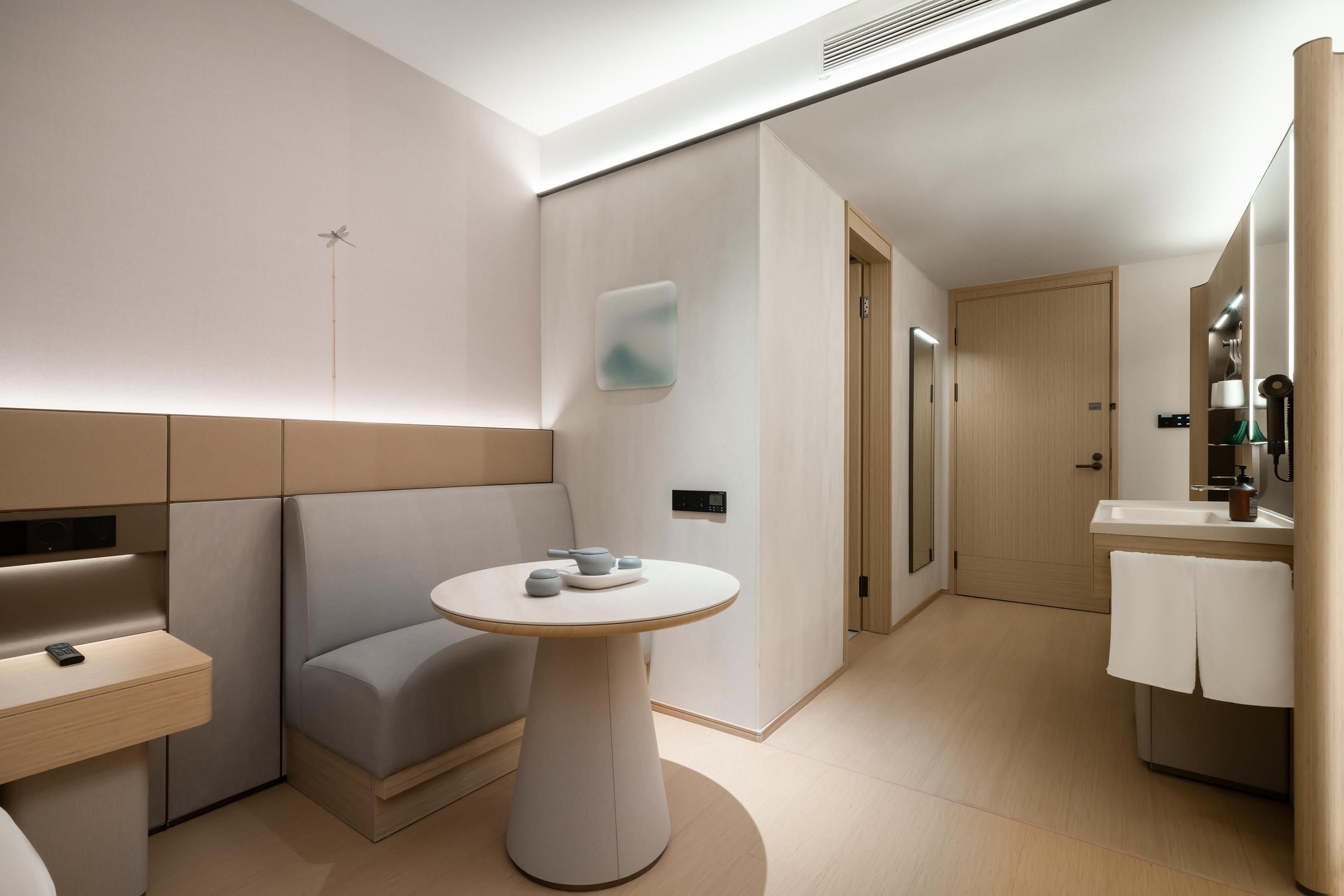 A modern hotel room with a minimalist design, featuring a sofa, a round table, a small bathroom with a sink and mirror, and a door leading to another room.