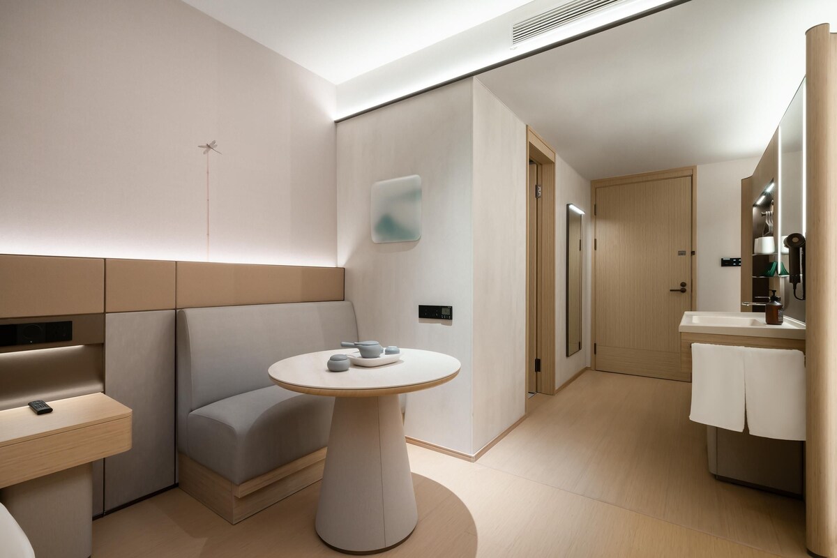 A modern hotel room with a minimalist design, featuring a sofa, a round table, a small bathroom with a sink and mirror, and a door leading to another room.