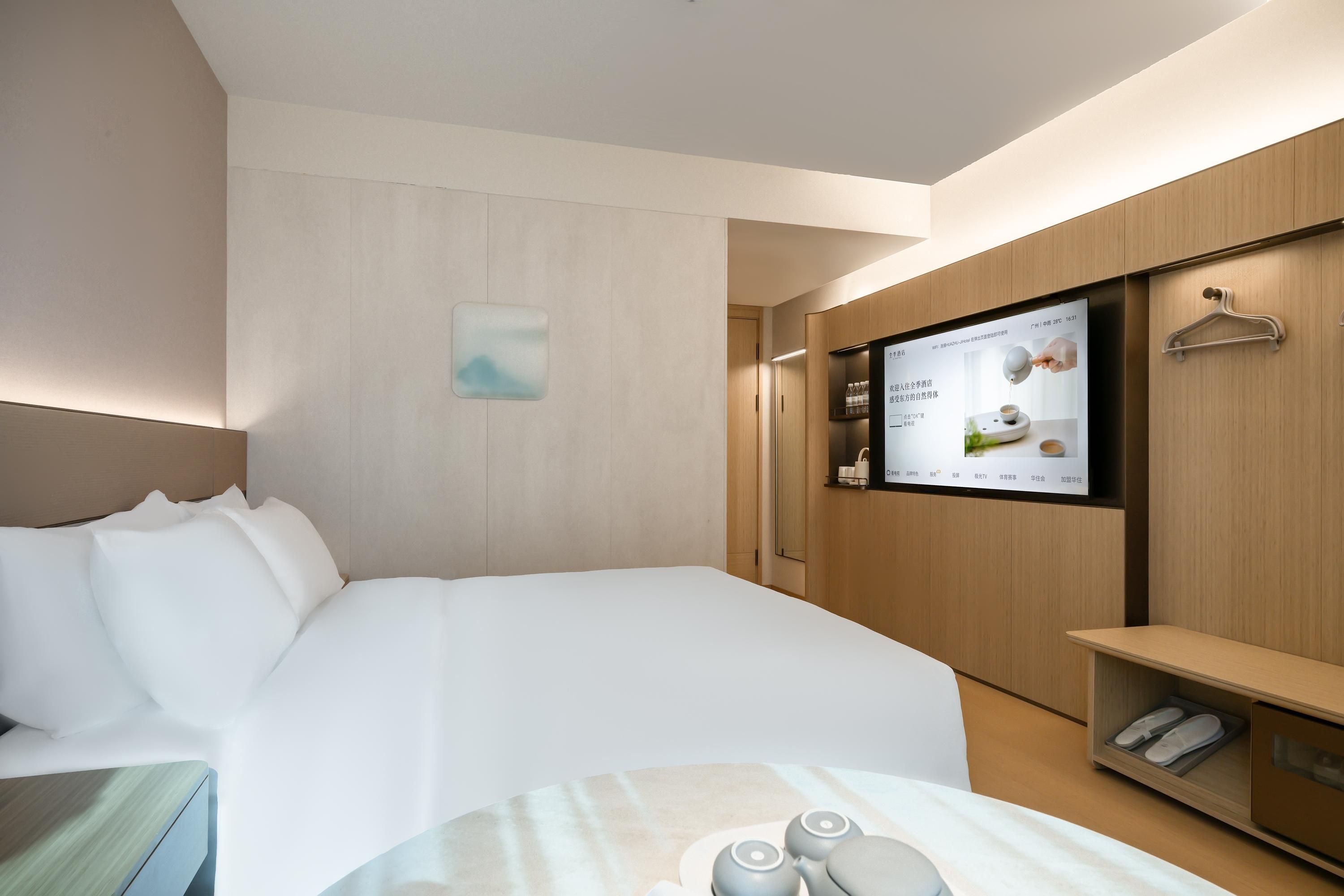 Quarto de hotel com cama, televisão fixada na parede, pequena mesa com bules e armário de madeira com sapateira.