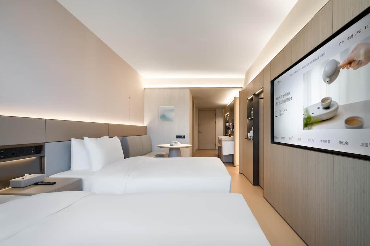 A modern hotel room with a large bed, bedside tables, a small round table, and a TV screen displaying an advertisement.