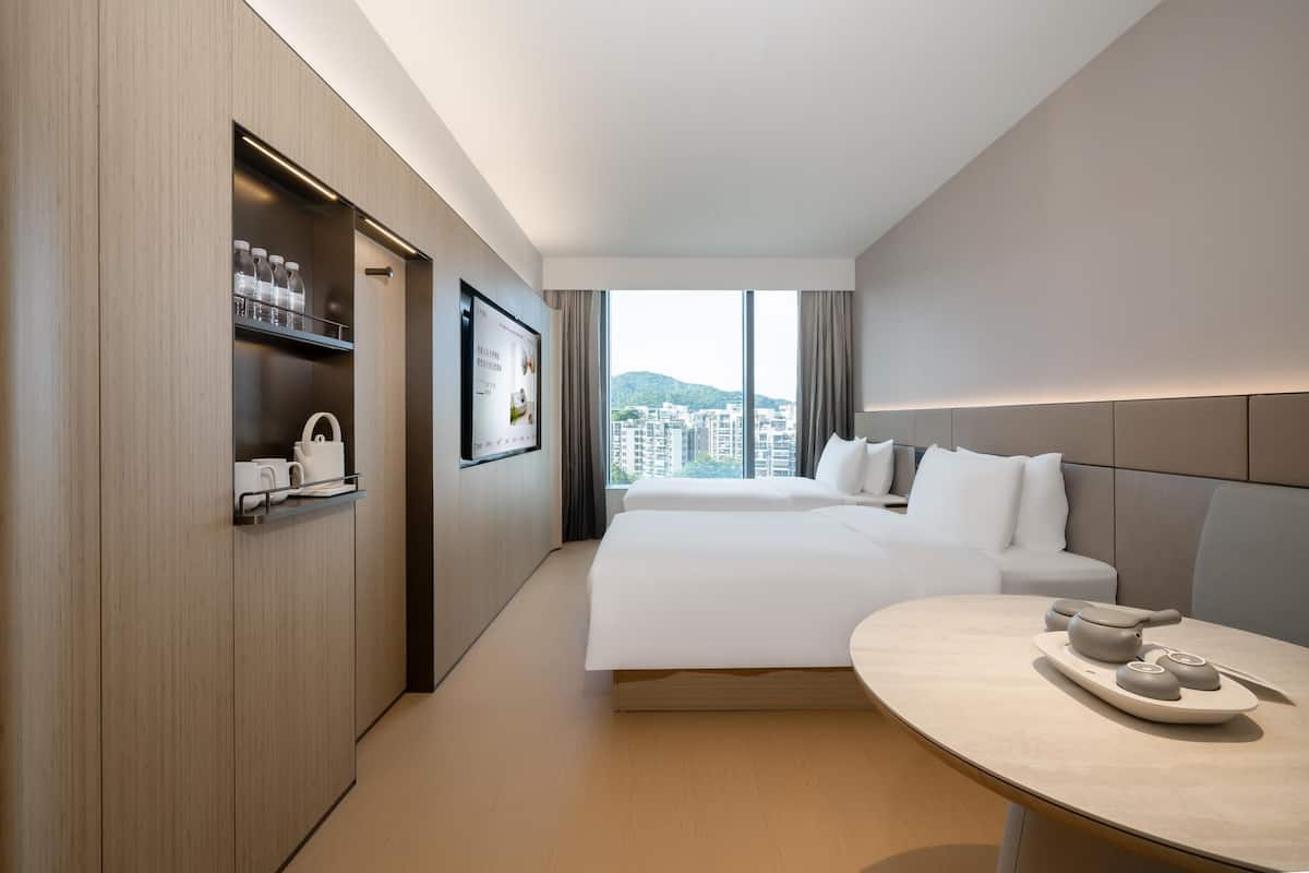 A modern hotel room with a large bed, a bedside table with a tray, a wardrobe with shelves, and a large window offering a city view.