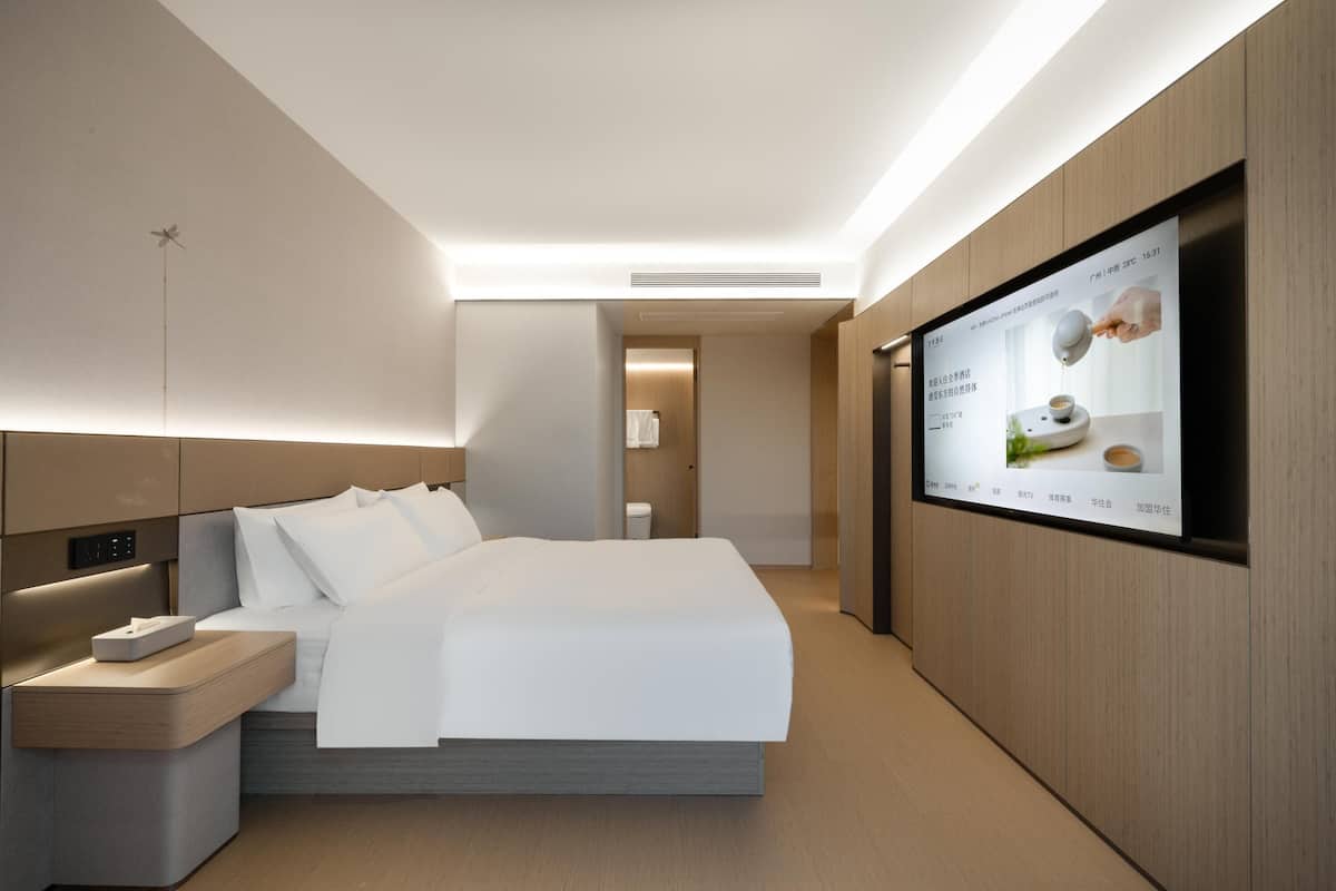 A modern hotel room with a large bed, a bedside table with a lamp, a TV mounted on the wall, and a wooden panel design.