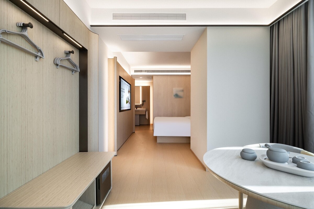 A modern hotel room with a wooden floor, a bed, a bedside table with a tray of items, and a wall-mounted TV.
