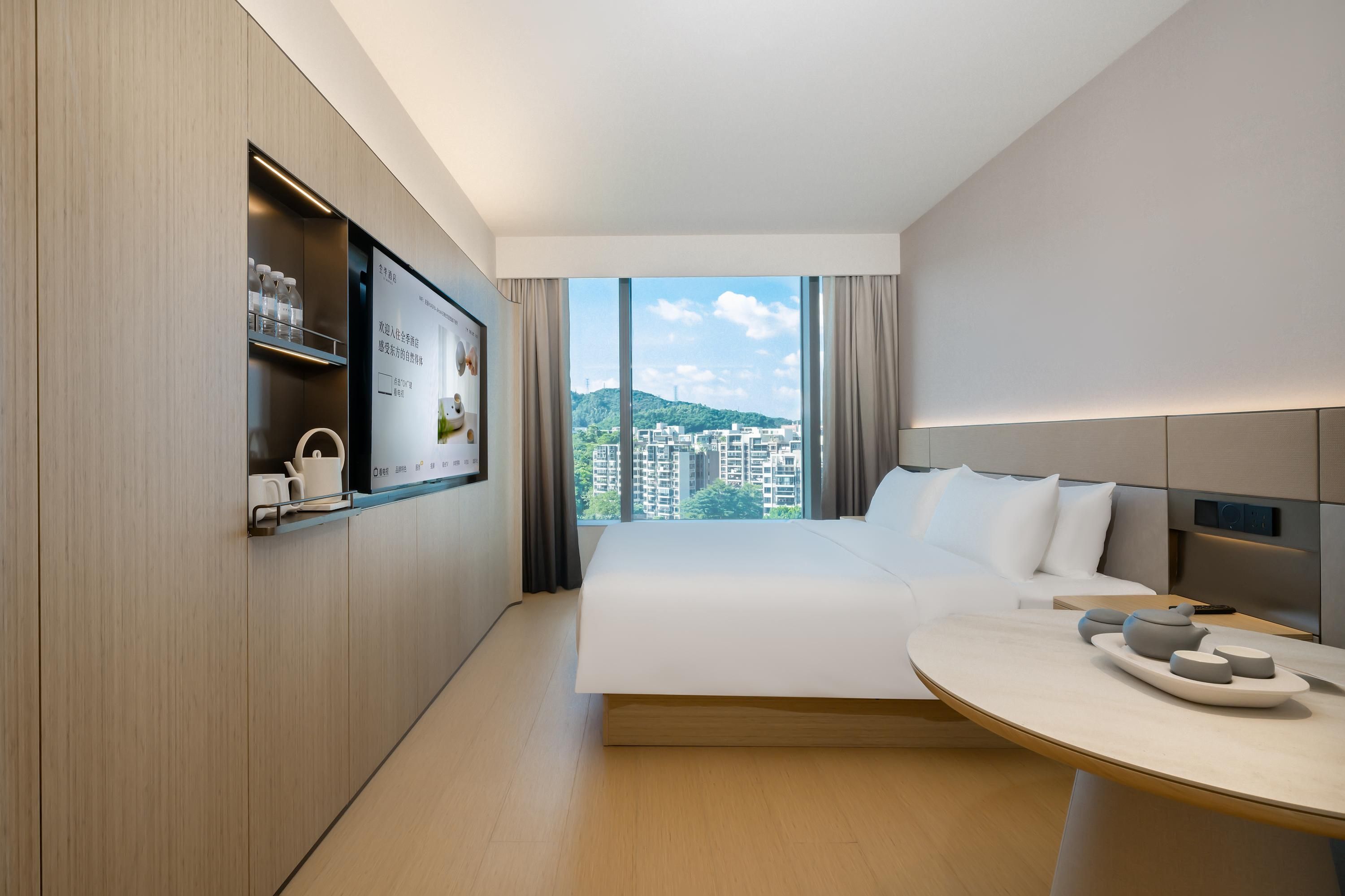 A modern hotel room with a large bed, a dining table, a TV mounted on the wall, and a view of the cityscape through large windows.