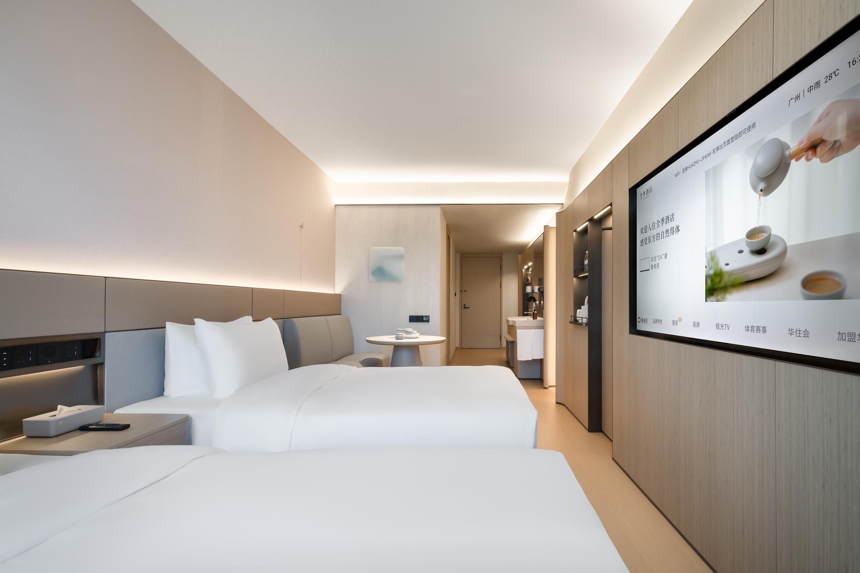 A modern hotel room with a large bed, bedside tables, a small table with tea set, and a wall-mounted TV displaying a tea advertisement.